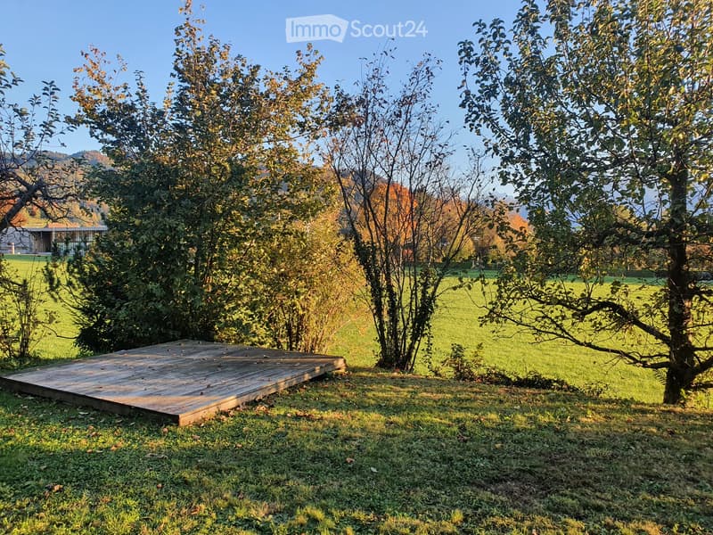 A wooden platform is situated in a grassy area surrounded by trees, with a modern house and open field in the background.