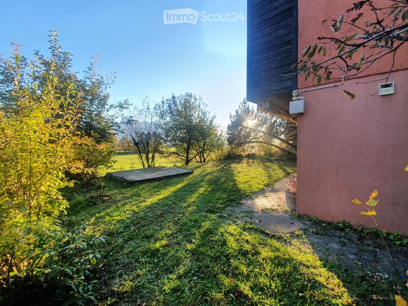Garden area with grass, trees, a wooden deck, and a pathway leading to a house with a balcony.
