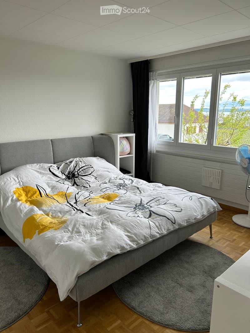 A modern bedroom with a grey bed, white and yellow floral duvet, nightstand, fan, radiator, and large windows with a scenic view.