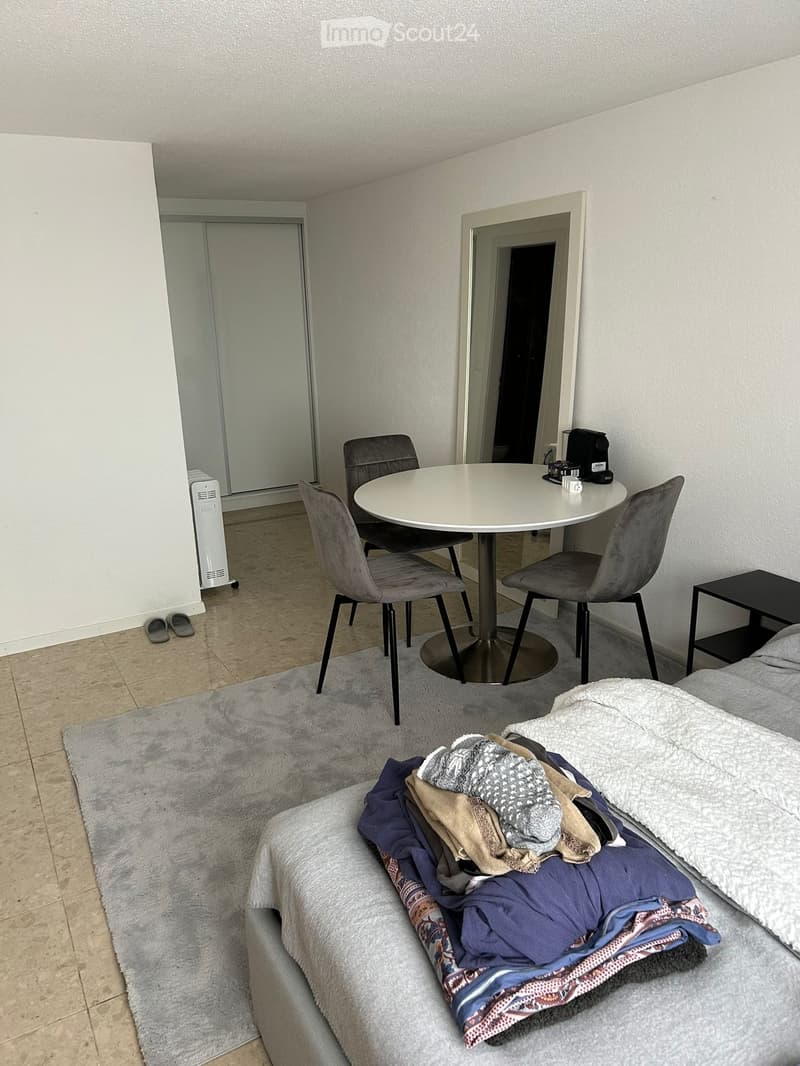 A small, furnished room with a bed, a round table with chairs, a nightstand, and a shower visible in the background.