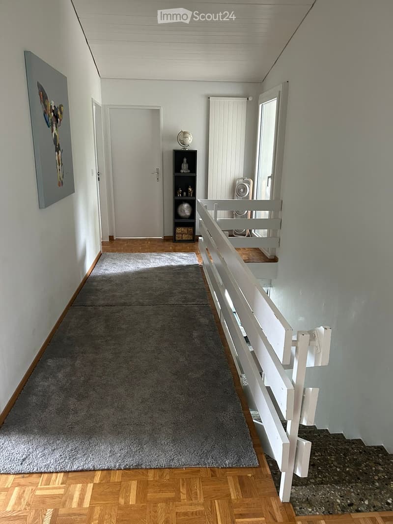 A well-lit hallway with wooden flooring, a gray carpeted area, white walls, and a staircase with a white railing. A small shelf with decorative items and a fan is visible near the staircase.
