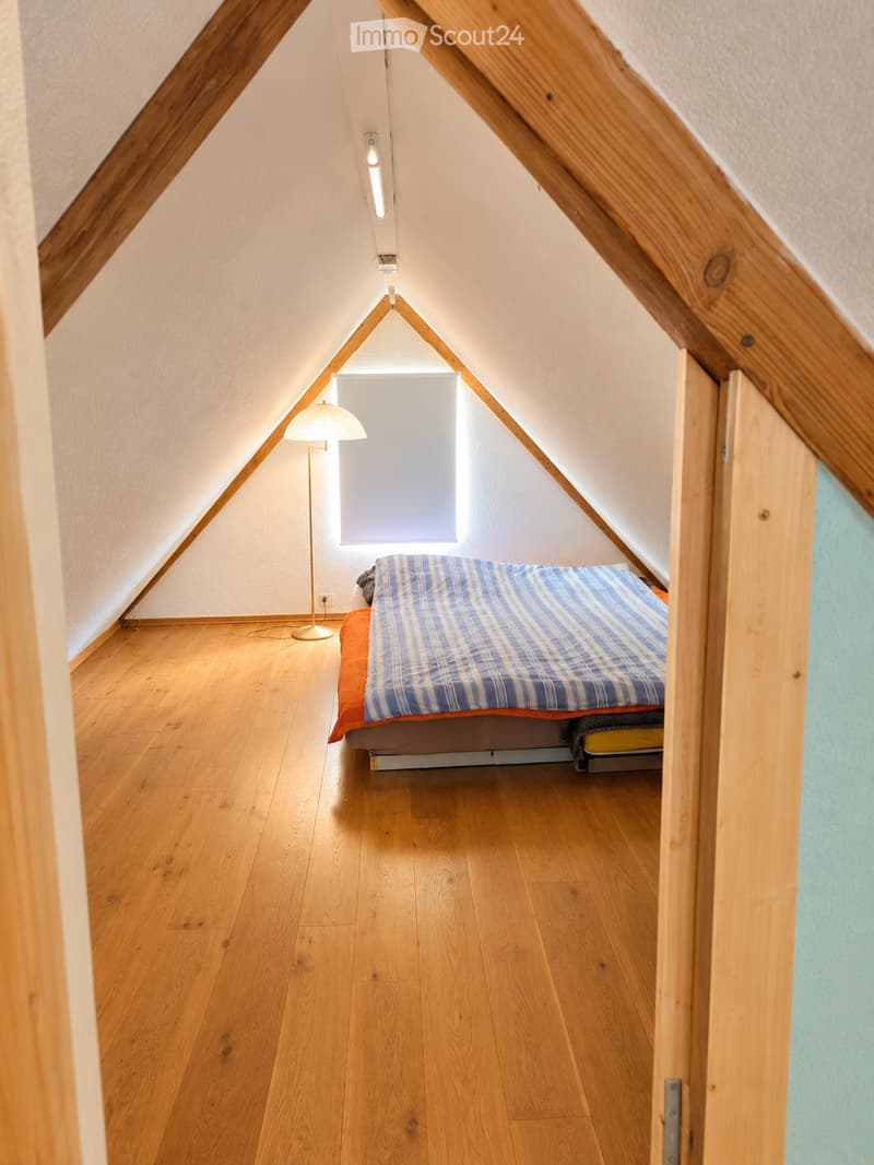 Cozy attic bedroom with wooden floors, slanted wooden ceiling beams, a single bed with blue and white striped bedding, and a floor lamp near the window.