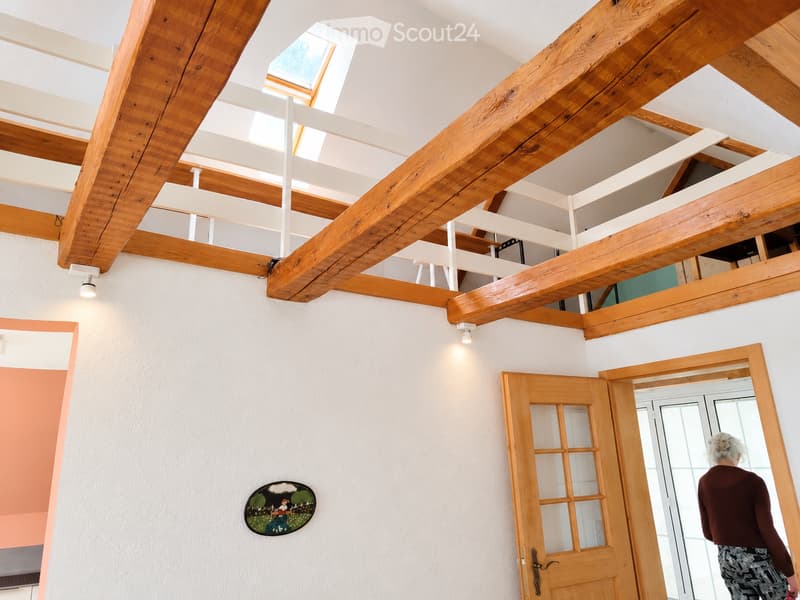 Bright hallway with exposed wooden beams and white walls, featuring a skylight, a round wall decoration, and a wooden door leading to another room.