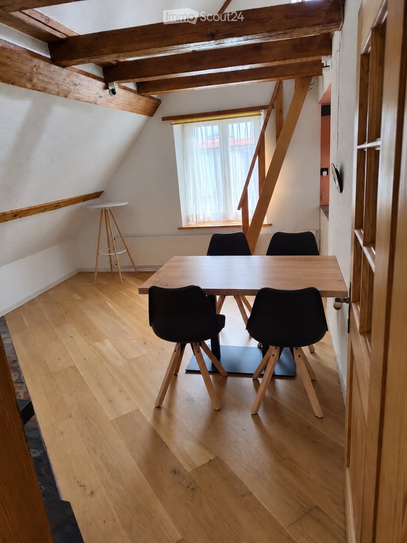Bright, wooden-floored room with exposed wooden beams, a large window with white curtains, a wooden table with four black chairs, and a small wooden stool near the window.