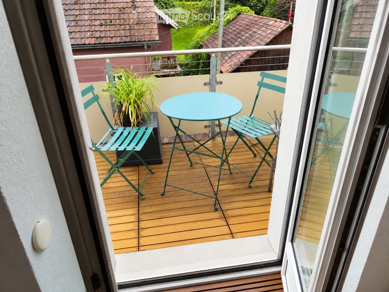 Balcony with wooden decking, turquoise metal table and chairs, potted plant, and railing overlooking a garden area.