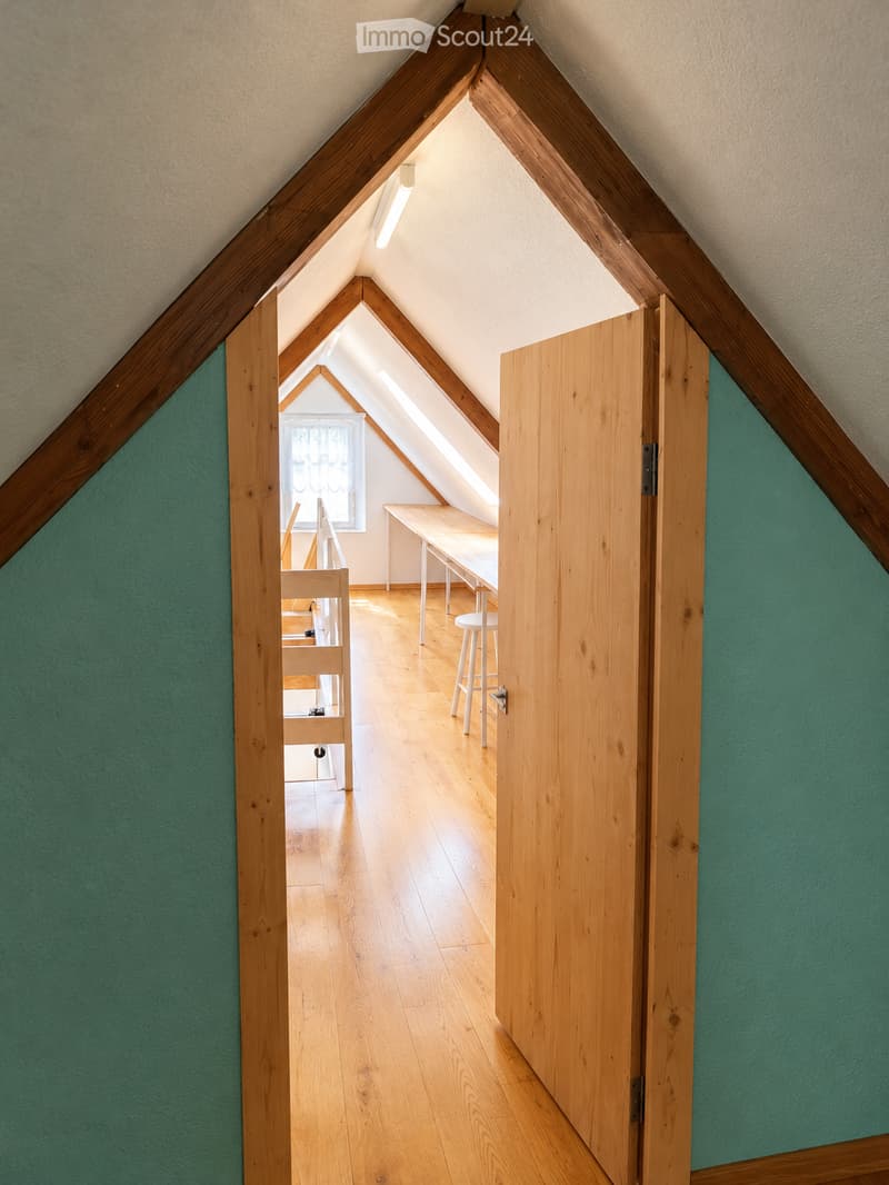 A narrow, slanted attic corridor with wooden beams and green walls, leading to a small workspace area with a desk, chair, and window.