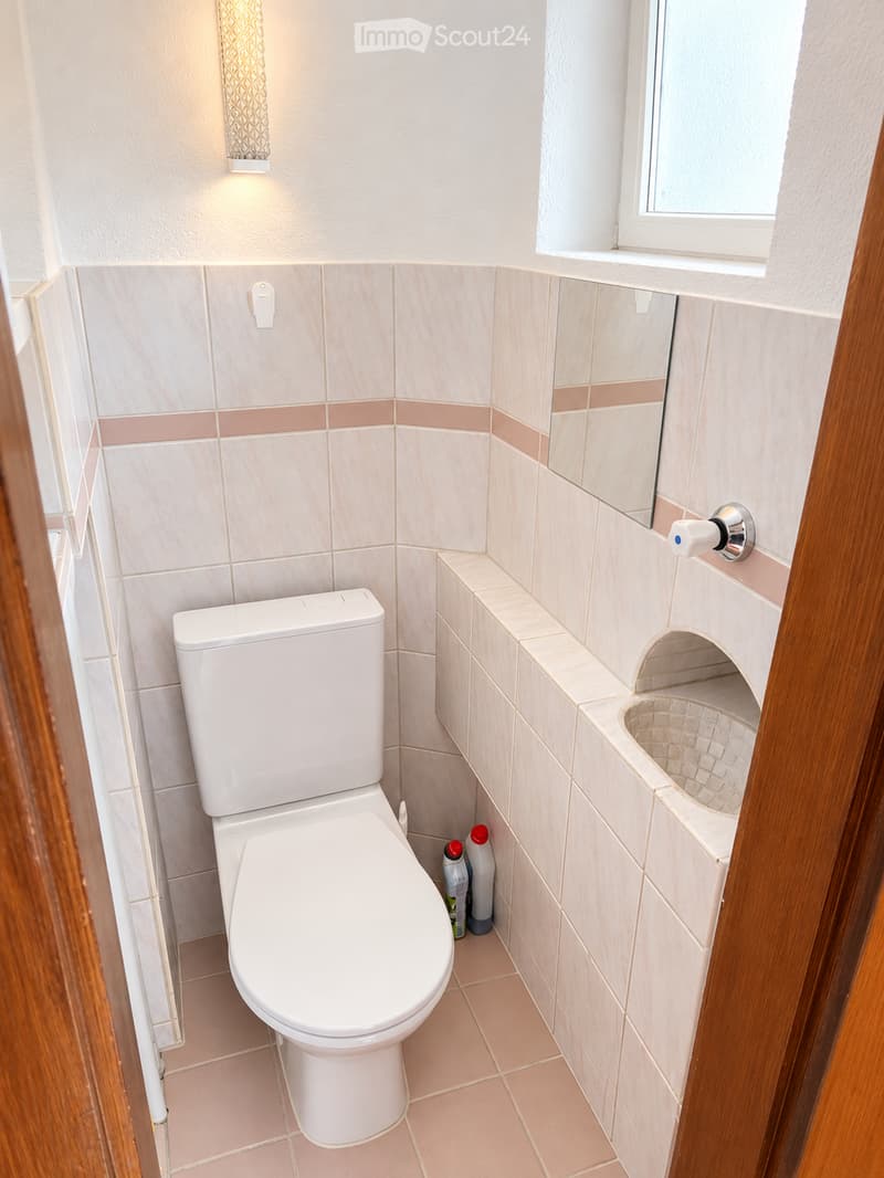 Modern bathroom with white toilet, tiled walls, a small sink, and cleaning supplies visible.