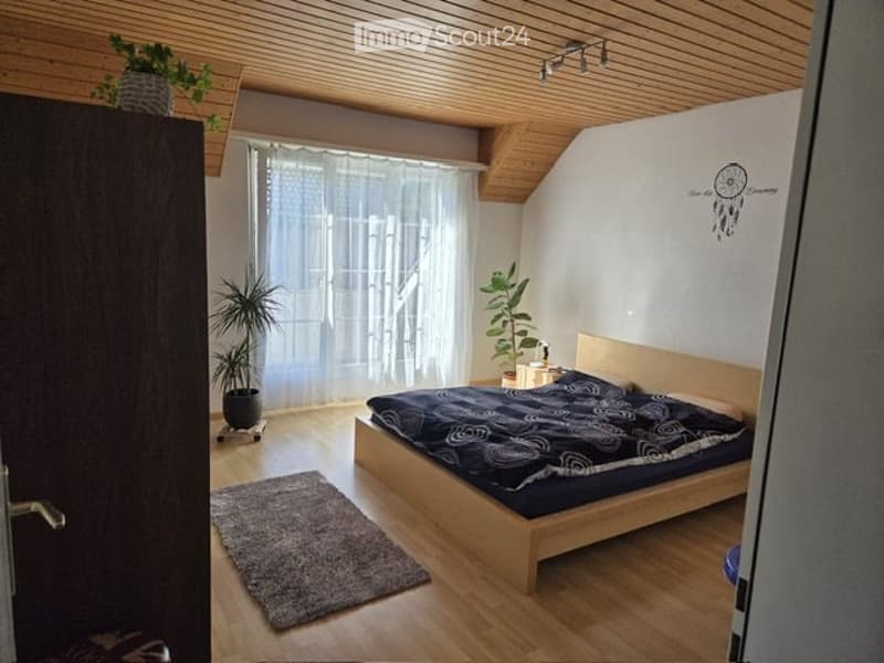 Cozy bedroom with wooden bed frame, dark blue bedding, nightstand, potted plants, and a large window with sheer curtains.