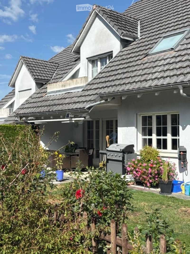 A well-maintained garden area in front of a modern house with a tiled roof and white exterior. The garden features a variety of flowering plants, shrubs, and a small patio area with outdoor furniture and a grill.