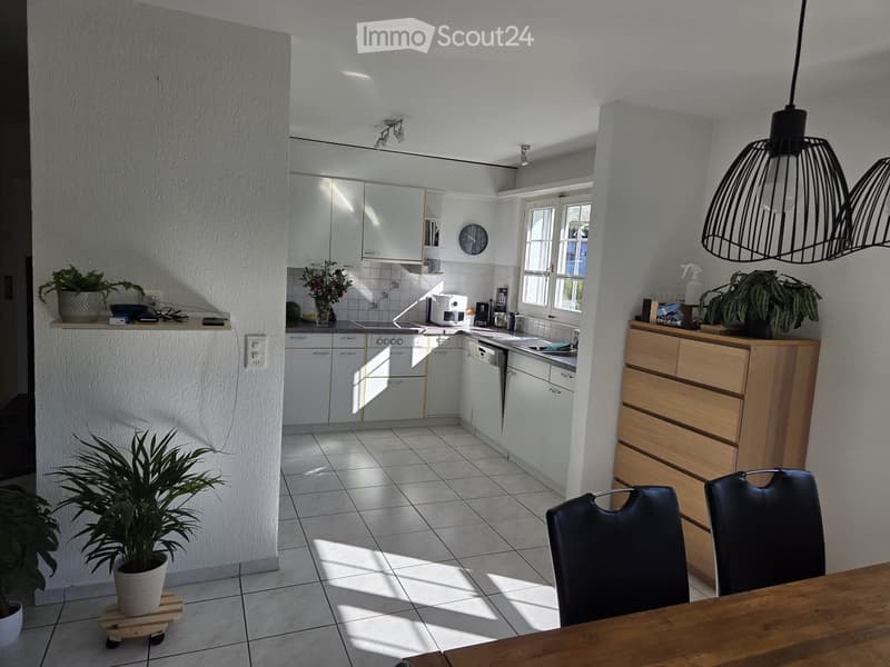 Bright, modern kitchen with white cabinets, a central island, and a dining area. Features include a wooden dining table, black chairs, potted plants, and various kitchen appliances.