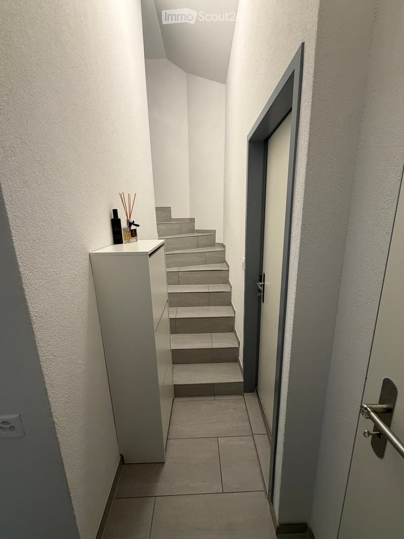A narrow hallway with tiled flooring, white walls, and a small table with candles on the left side. A staircase leads upwards, and a door is visible on the right.