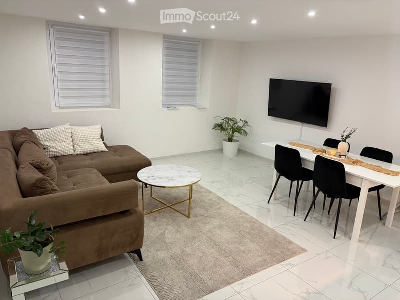 Modern living room with a brown sectional sofa, white coffee table, white dining table with four black chairs, and a flat-screen TV mounted on the wall. The room features white walls, large windows with blinds, and a light-colored rug.