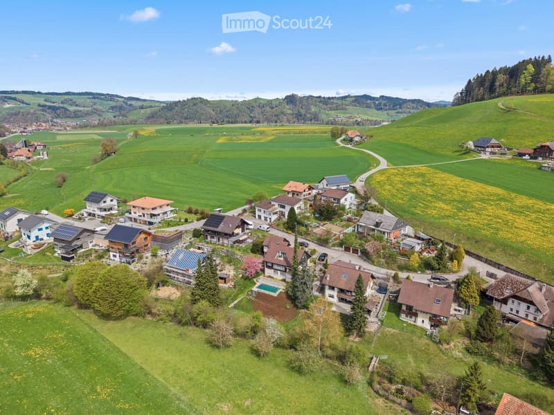 Aerial view of a rural village with multiple houses, green fields, and surrounding hills.