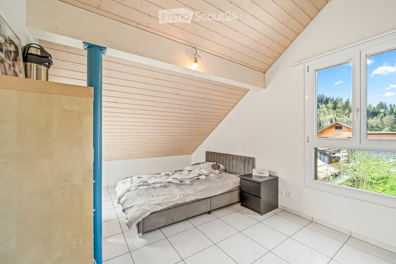 Cozy bedroom with a modern bed, nightstand, and large windows offering scenic views. The room features a slanted wooden ceiling and tiled flooring.