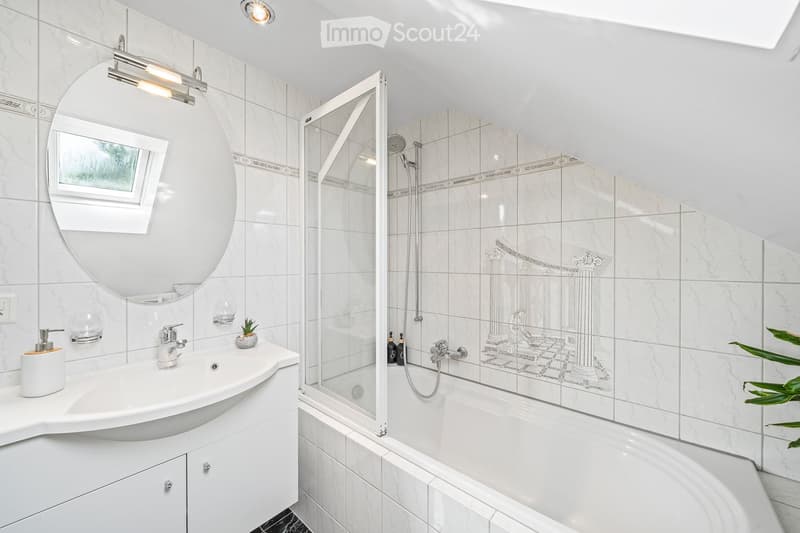 Modern bathroom with white marble tiles, a large oval mirror, a sleek sink with a modern faucet, a bathtub with a glass shower enclosure, and decorative plant on the counter.