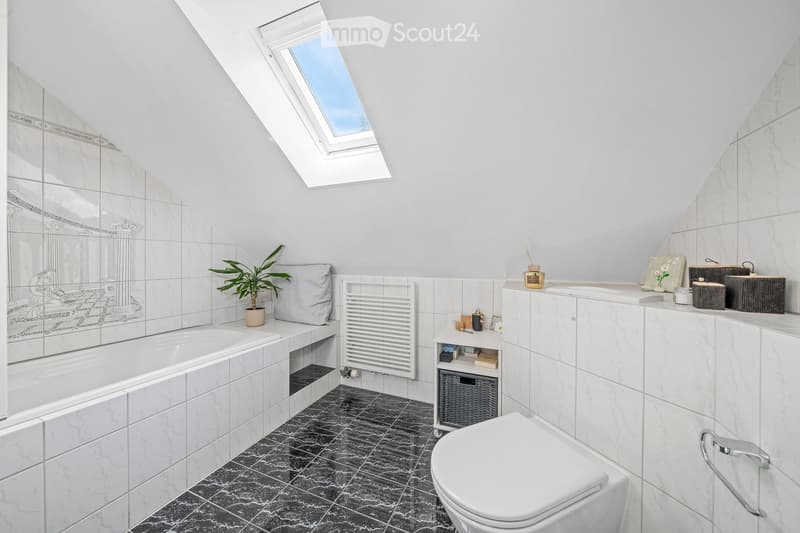 Modern bathroom with a bathtub, toilet, sink, and a large mirror. Features marble tiles, a skylight window, and decorative items on the sink counter.