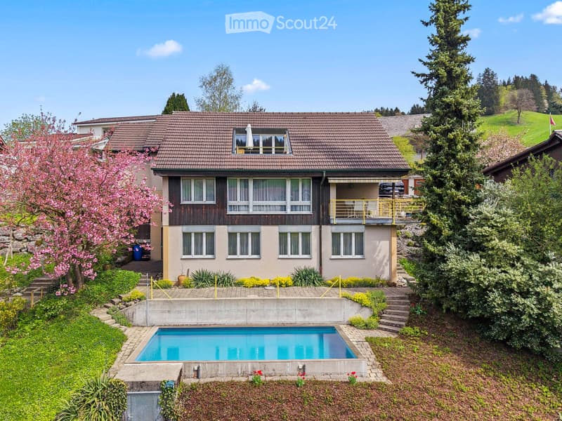 Two-story house with a tiled roof, multiple windows, and a balcony. The property includes a swimming pool in the garden, surrounded by greenery and flowers.