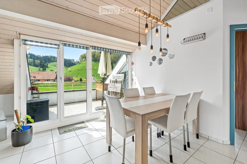 Bright kitchen with white tiled flooring, wooden dining table surrounded by white chairs, large windows offering a scenic countryside view, and a potted plant near the window.