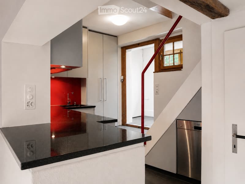 Modern kitchen with white cabinets, black countertops, red backsplash, and stainless steel dishwasher. Features a sleek design with a staircase leading upwards.