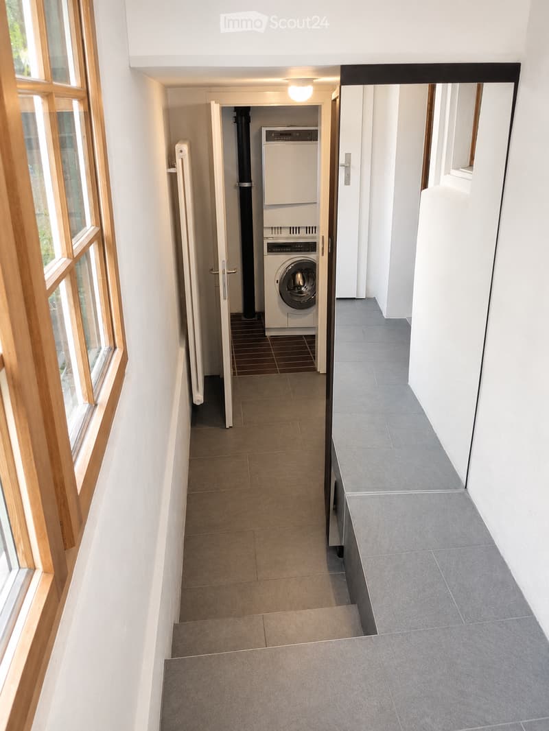 A well-lit hallway with tiled flooring, white walls, and large wooden-framed windows on one side. A door leads to a room containing a washing machine and dryer.
