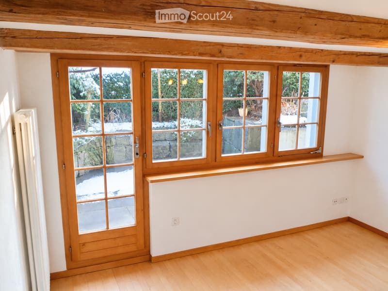 Empty room with wooden flooring, white walls, large wooden-framed windows, and a radiator. No furniture or personal items are present.