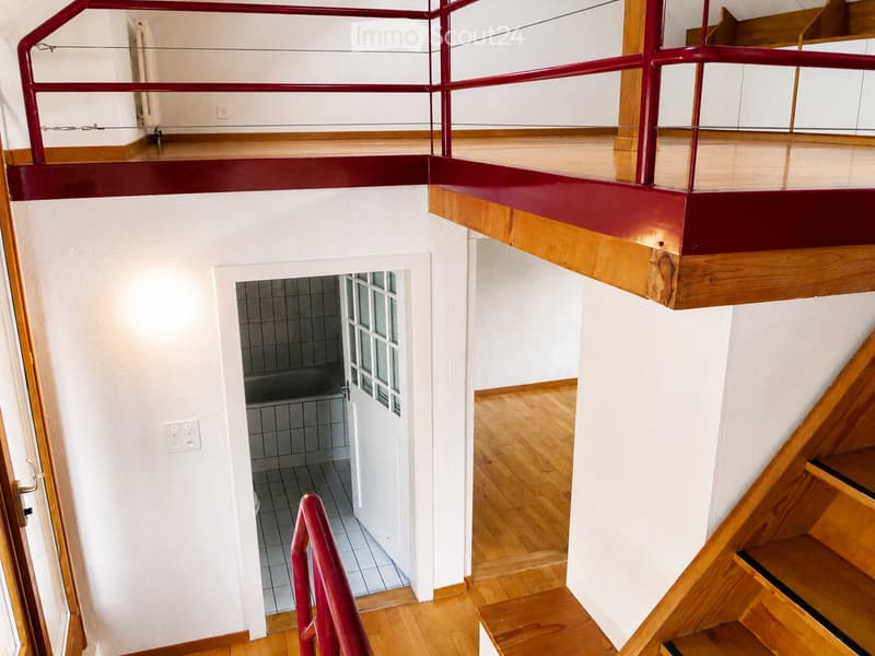 View of a modern interior corridor with wooden flooring, white walls, and a staircase leading upwards. A small tiled bathroom is visible through an open doorway. The corridor features a red metal railing and a radiator on the wall.