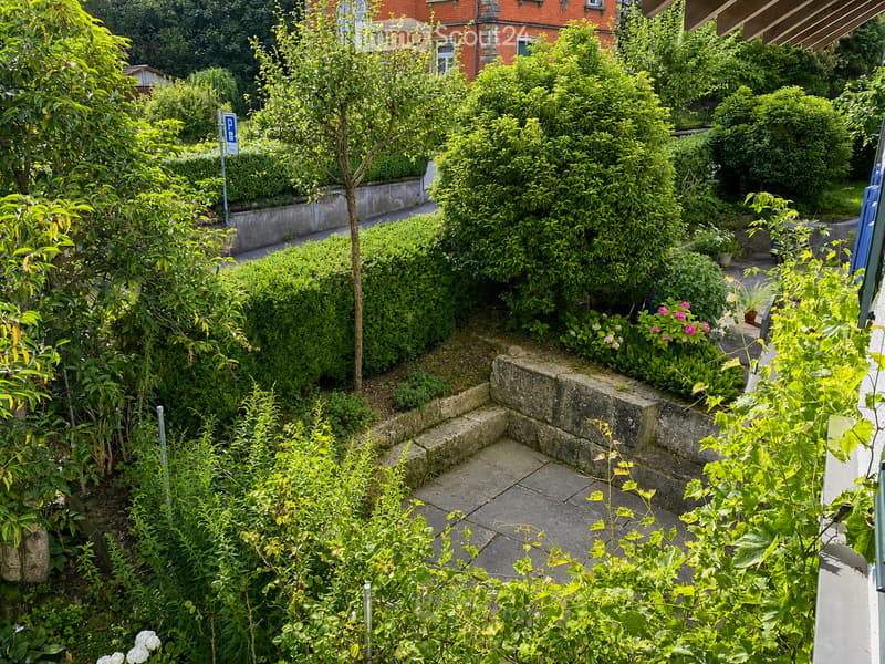 A well-maintained garden with various green plants, shrubs, and flowers arranged on a stone-paved pathway, with a brick building and parking sign visible in the background.