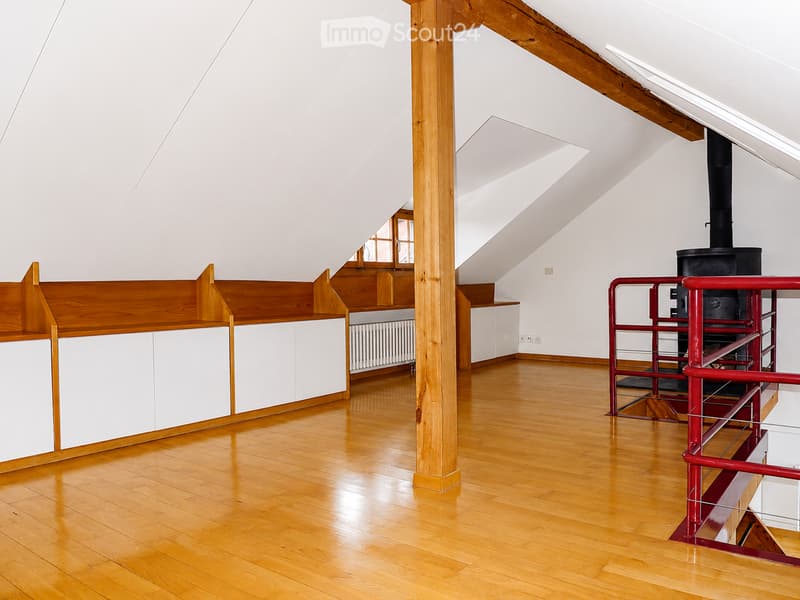 Bright, empty attic room with wooden floors, white walls, exposed wooden beams, and a staircase leading to another level.