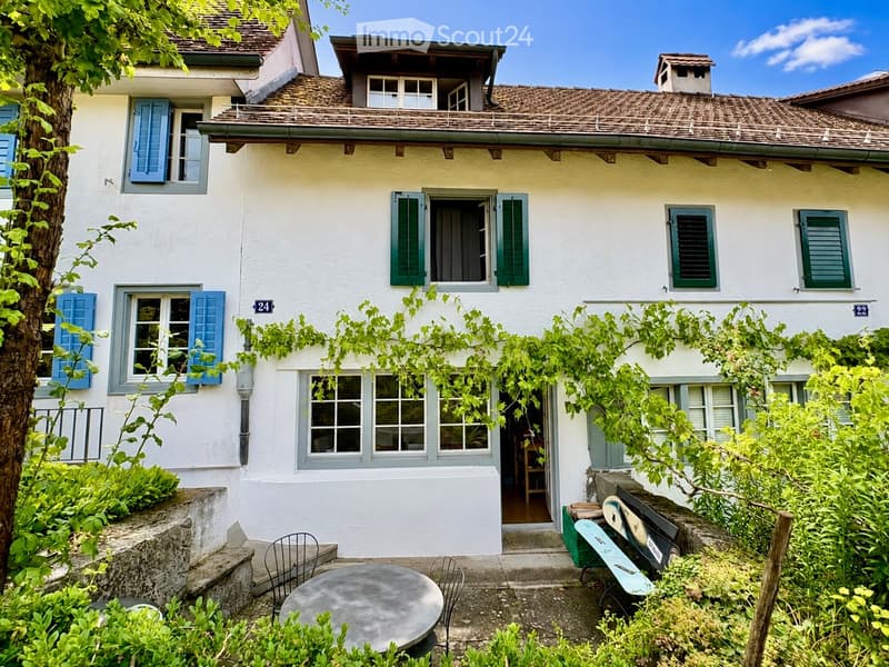 Two-story house with white walls, blue shutters, and a tiled roof. The house has multiple windows and a small balcony on the upper floor. The entrance is surrounded by greenery, with outdoor seating including a round table and chairs.