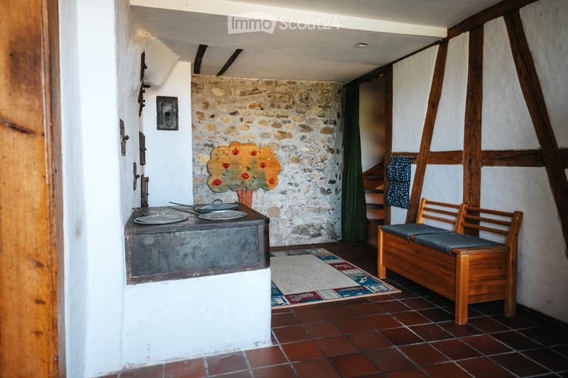 A rustic room with exposed wooden beams, stone walls, a wooden bench with blue cushions, a small table with plates, and a colorful painting of a tree on the wall.