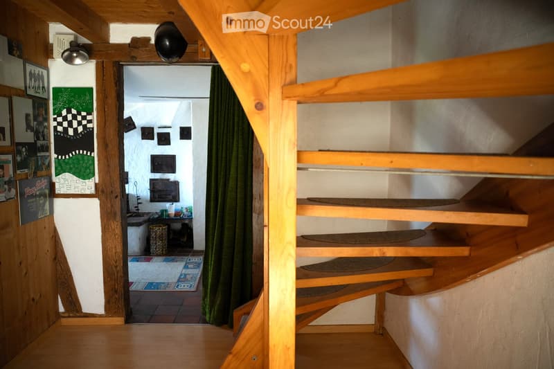 A narrow wooden hallway with exposed beams and a staircase leading to a small kitchen area. The kitchen features a sink, countertop, and various small appliances.