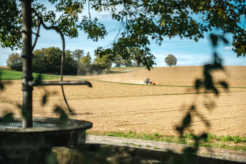 A wide, open field with a tractor moving across the landscape, surrounded by trees and a clear blue sky.