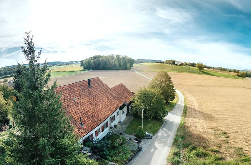 A two-story house with a red-tiled roof, surrounded by trees and open fields. The house has a small porch area with outdoor furniture and is situated in a rural landscape with agricultural fields and distant hills.