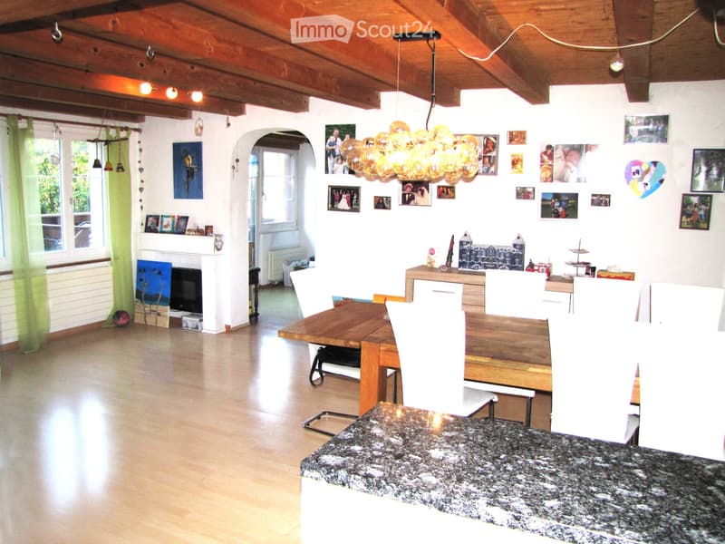 A well-lit living and dining area with wooden floors, a large wooden dining table surrounded by white chairs, a fireplace with a TV, and a balloon light fixture hanging from the ceiling. The walls are decorated with framed photos and artwork.