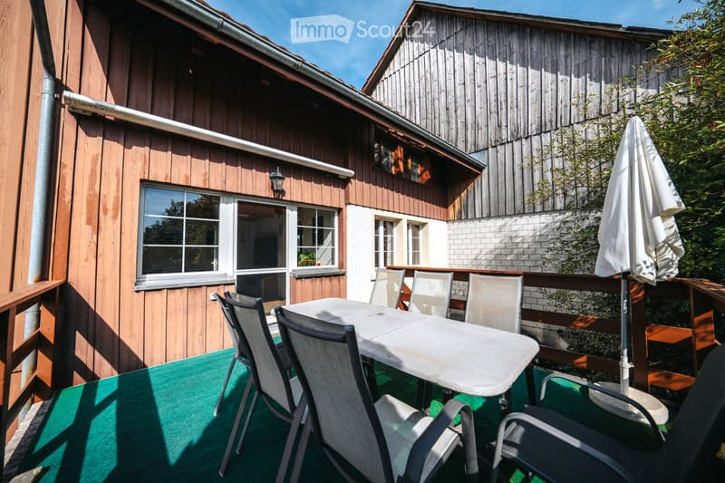 Balcony with a green artificial turf floor, a white table, and several grey chairs. A large umbrella is positioned near the railing, and the background shows part of a wooden house with windows and a lantern-style light fixture.