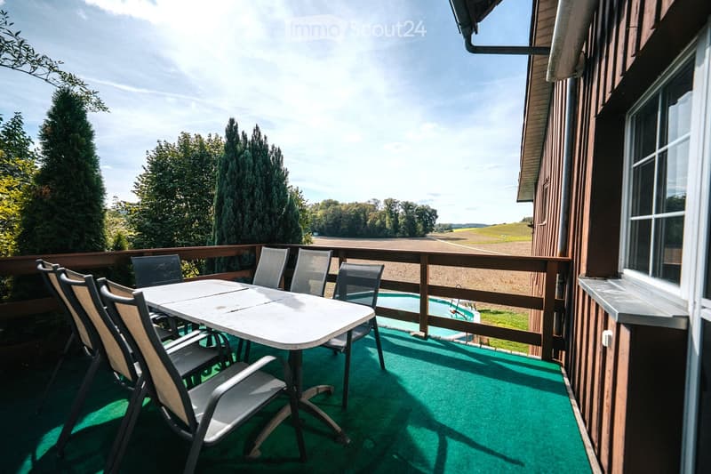 Balcony with green artificial turf, a white table, and several chairs, overlooking a rural landscape with trees and fields.