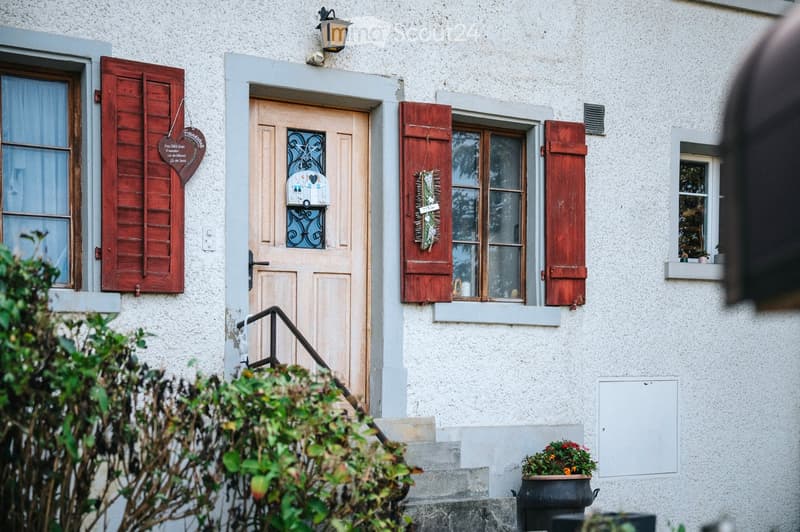 Front entrance of a house with a light-colored door, decorative shutters, and a small staircase leading up to it. The house has a textured white facade, and there are potted plants near the entrance.