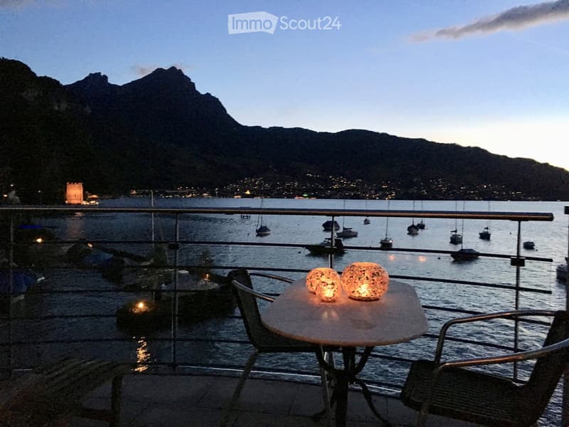 Balcony with a round table, two chairs, and decorative lights, overlooking a harbor with boats and a mountainous backdrop.