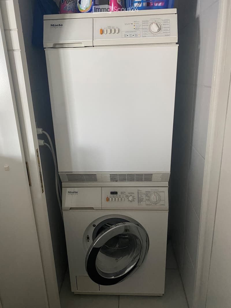 A compact bathroom with a stacked Miele washing machine and dishwasher unit, white tiled walls, and a small shelf holding cleaning supplies above the appliances.