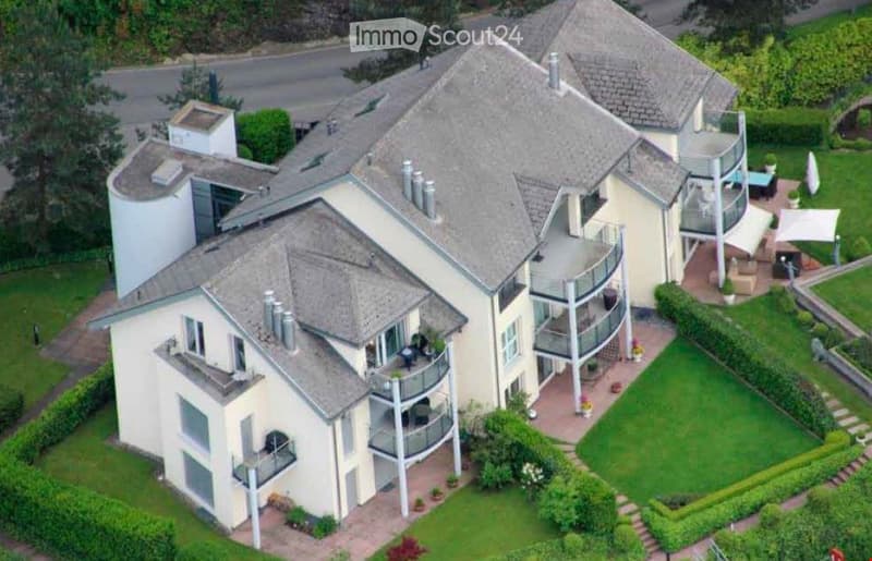 A large, modern multi-story house with multiple balconies, a well-maintained garden, and a patio area with outdoor furniture.