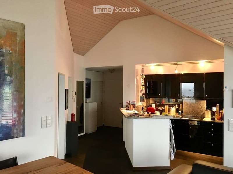 A modern kitchen with black cabinets, a white island, and a dining area with a wooden table and chairs. The space features a slanted wooden ceiling and a large abstract painting on the wall.