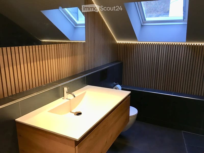 Modern bathroom with a sleek wooden vanity, integrated sink, chrome faucet, large mirror, and a toilet. The walls feature vertical wooden paneling and built-in LED lighting, with skylights providing natural light.
