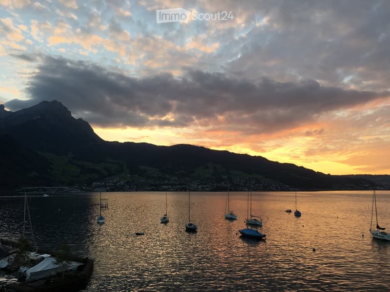 A serene lakeside scene at sunset with sailboats anchored in calm water, surrounded by mountains and a town in the distance.