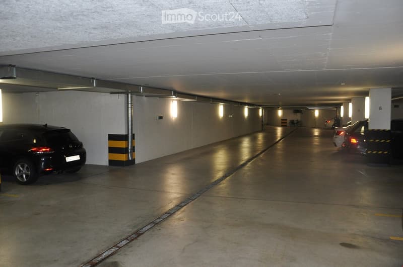 Underground parking area with several parked cars, concrete floors, and white walls with vertical lighting fixtures.