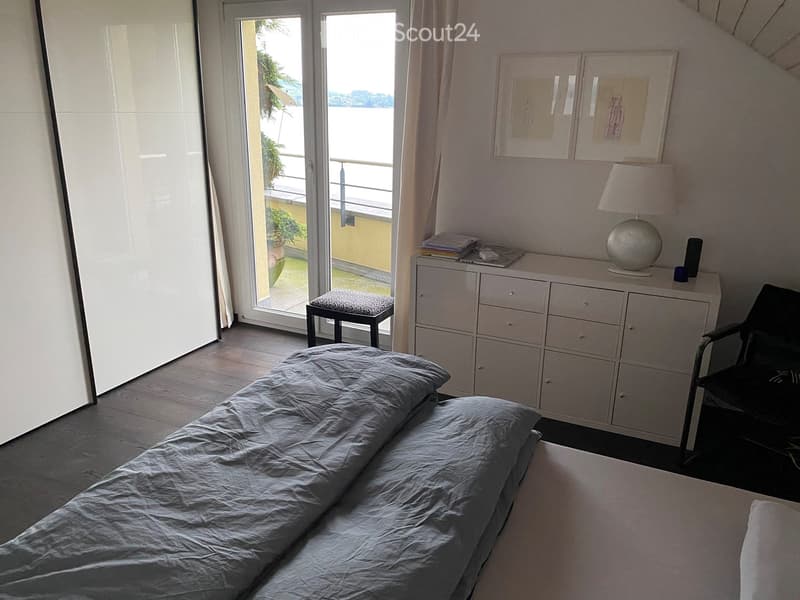 Bright bedroom with a large bed, a white cabinet, a lamp, a chair, and a small stool near the balcony door offering a view.
