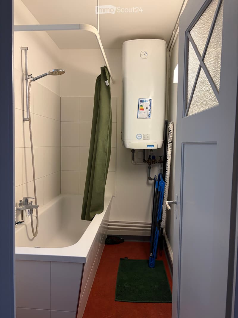 Compact bathroom with a bathtub, showerhead, water heater, and a green bathrobe hanging on the side.