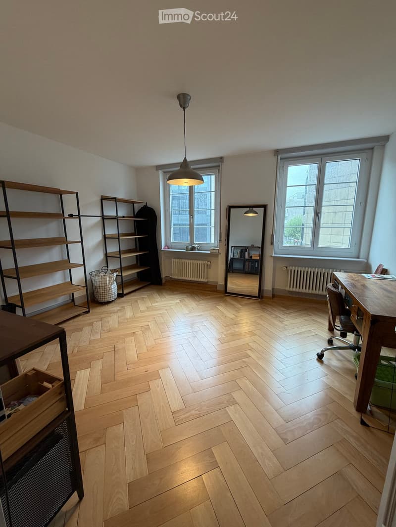 Bright room with herringbone wooden flooring, large windows, a hanging lamp, a desk with a chair, shelves, and a mirror.