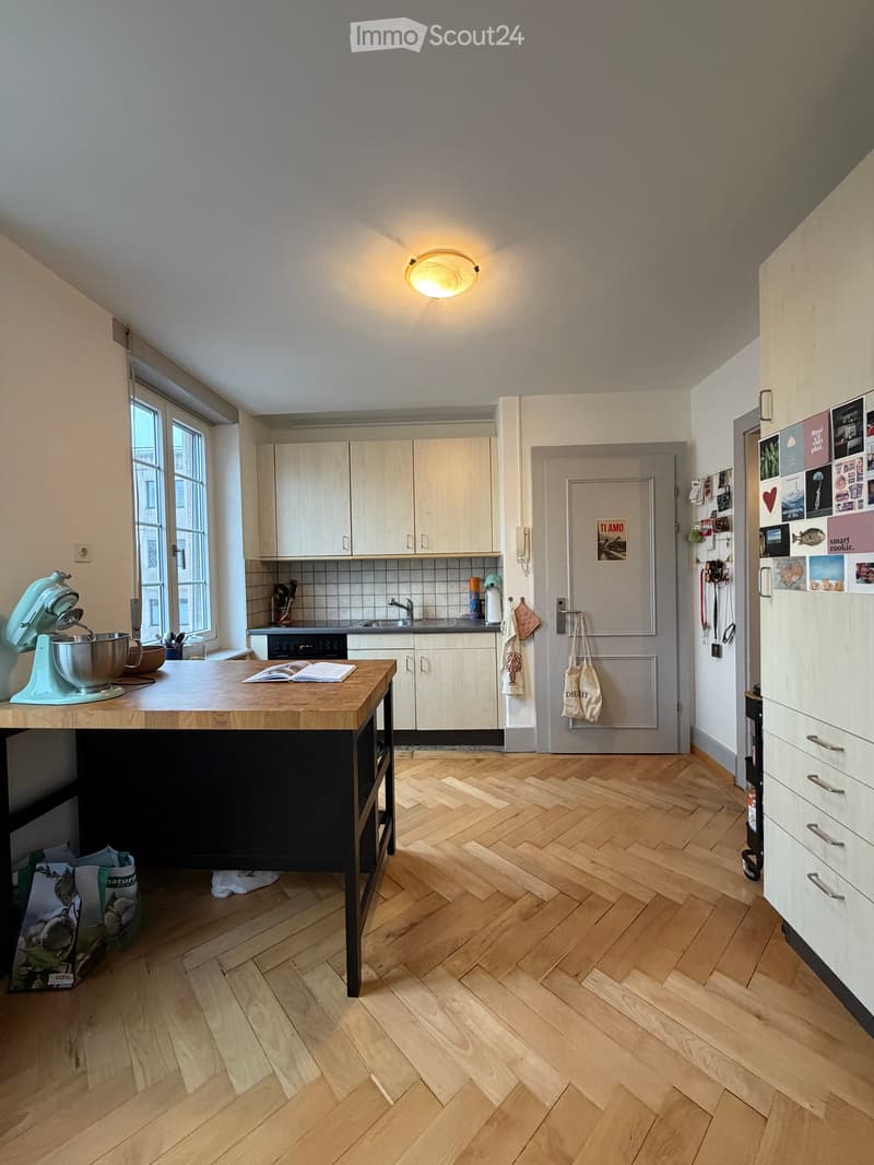 Modern kitchen with light wooden cabinets, herringbone wooden flooring, and a central island with a mixer and bowl. The kitchen features a sink, oven, and refrigerator with various magnets and notes.