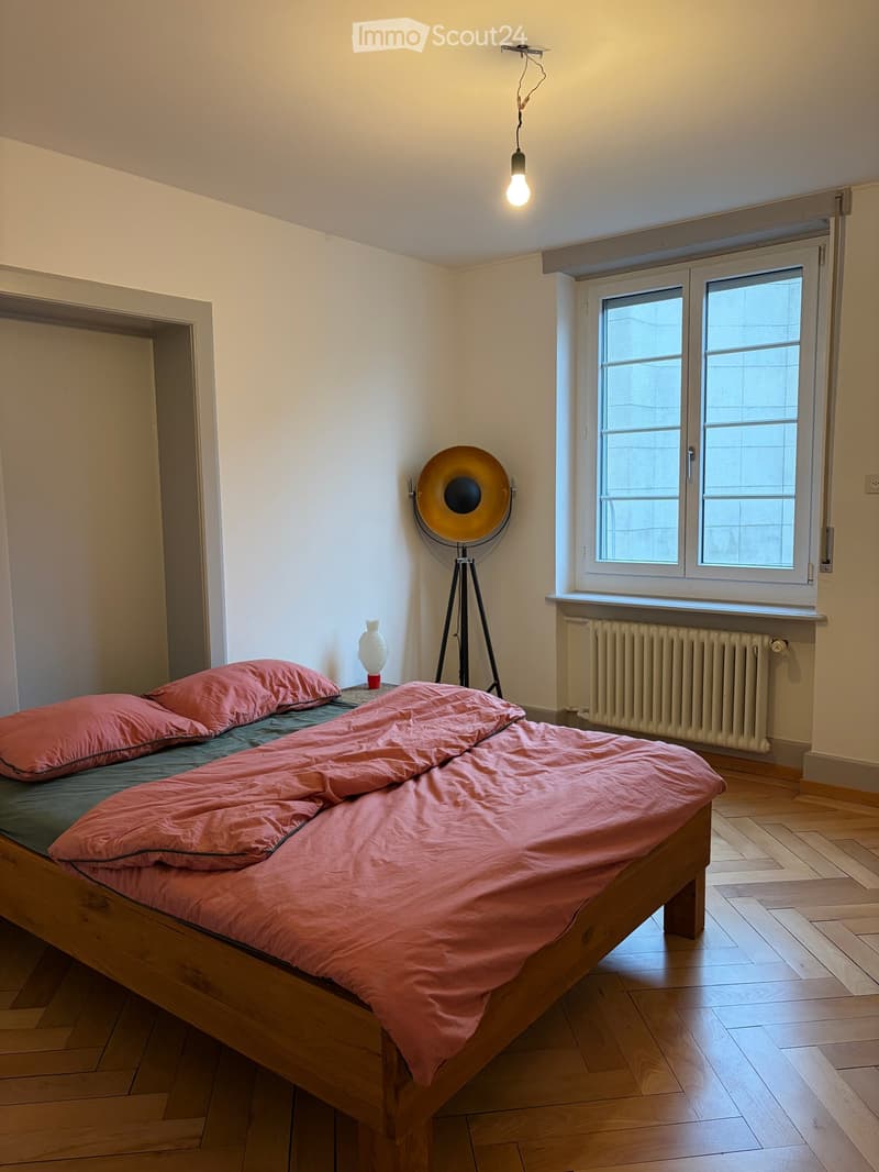 A small, well-lit bedroom with a wooden bed, pink bedding, a floor lamp, and a vase on a nightstand.