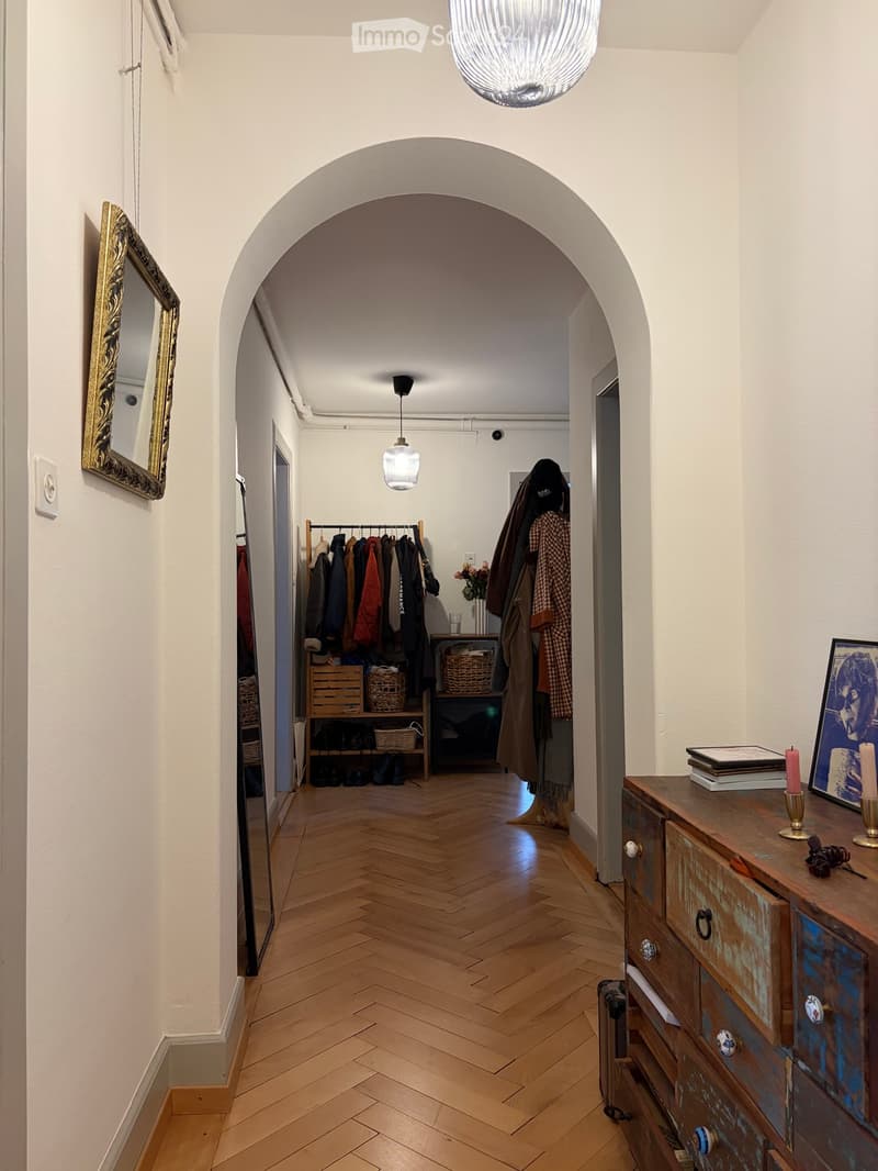 A well-lit hallway with wooden herringbone flooring, white walls, and a large arched doorway leading to another room. Features include a coat rack with jackets, a small table with a vase of flowers, and a dresser with books and candles on top.